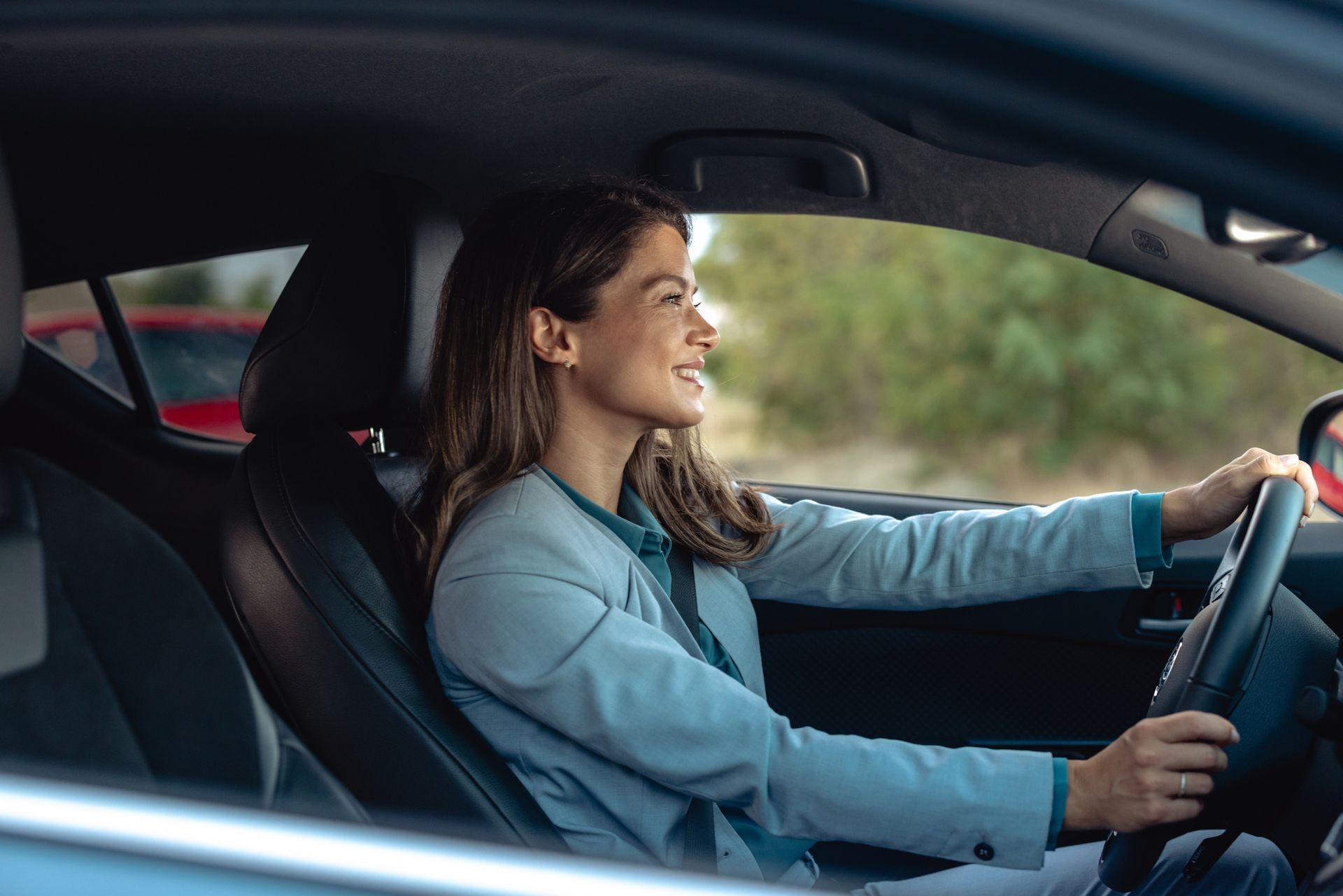 Woman smiling while driving a car, holding the steering wheel. She's wearing a blue blazer and seatbelt.