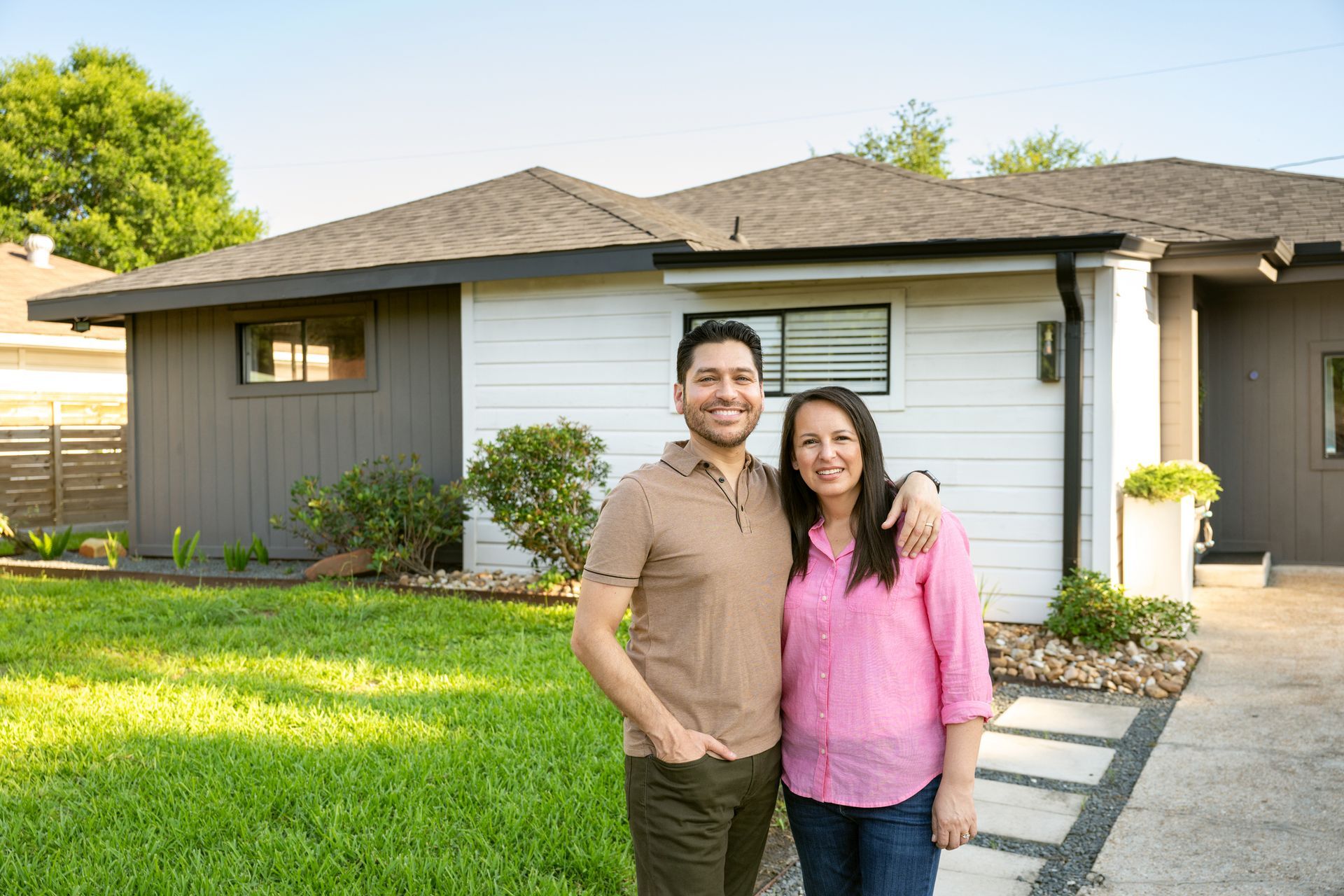 Couple standing in front of renovated house; man with brown shirt, woman with pink shirt, both smiling.