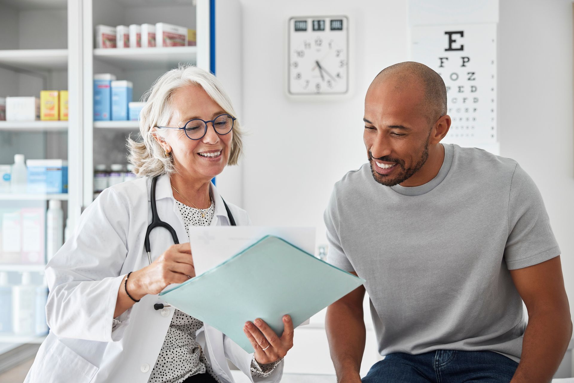 Doctor smiling, showing a file to a patient in an exam room. White coat, stethoscope.