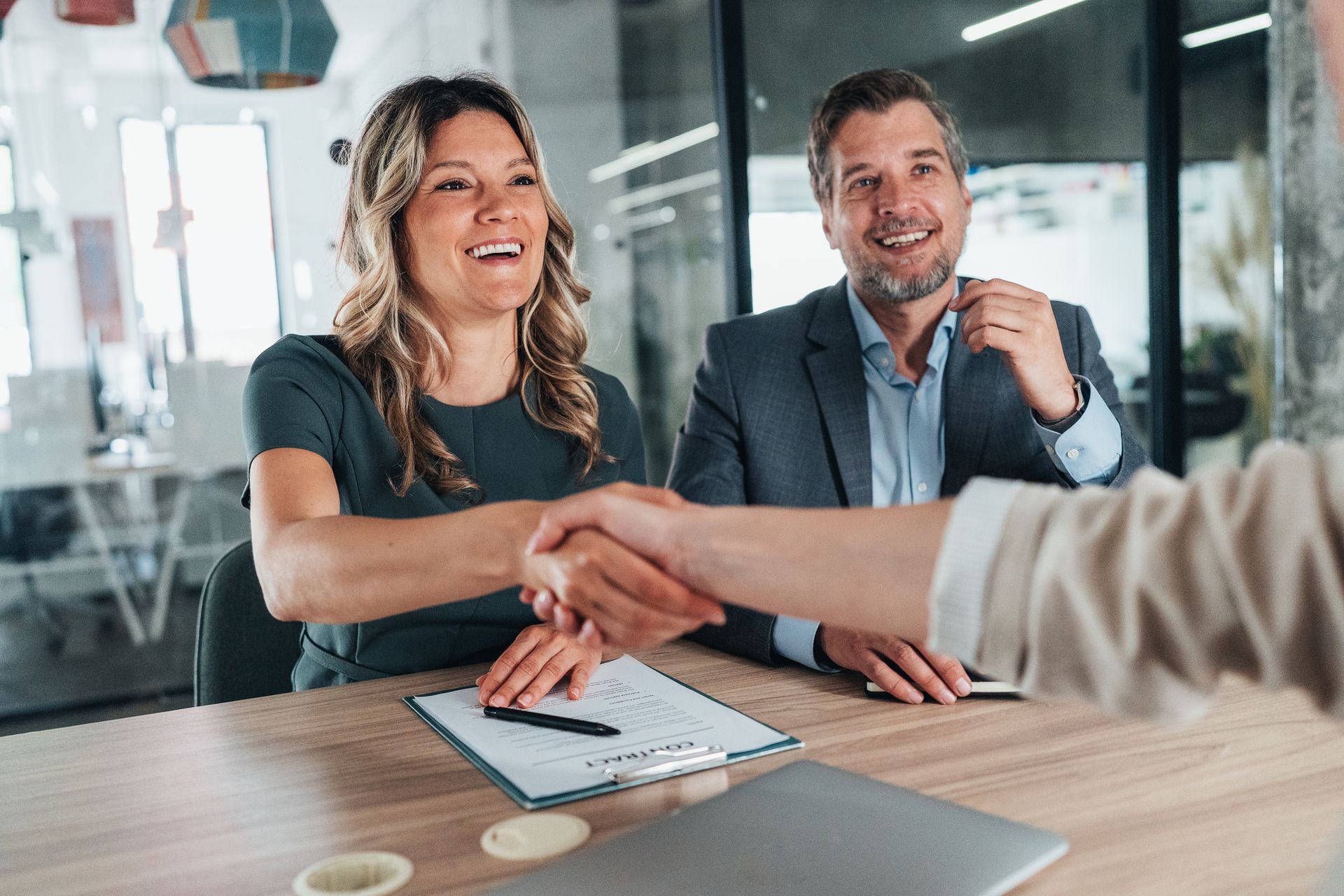 Couple shaking hands with another person at a table, smiling in an office.
