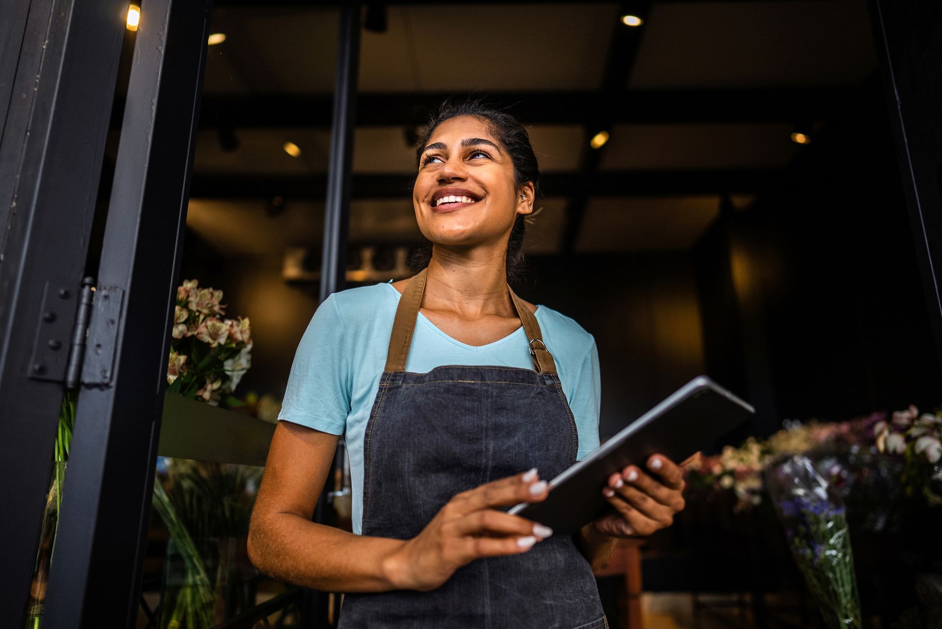 Woman in apron, holding a tablet, smiles while standing in a doorway of a flower shop.