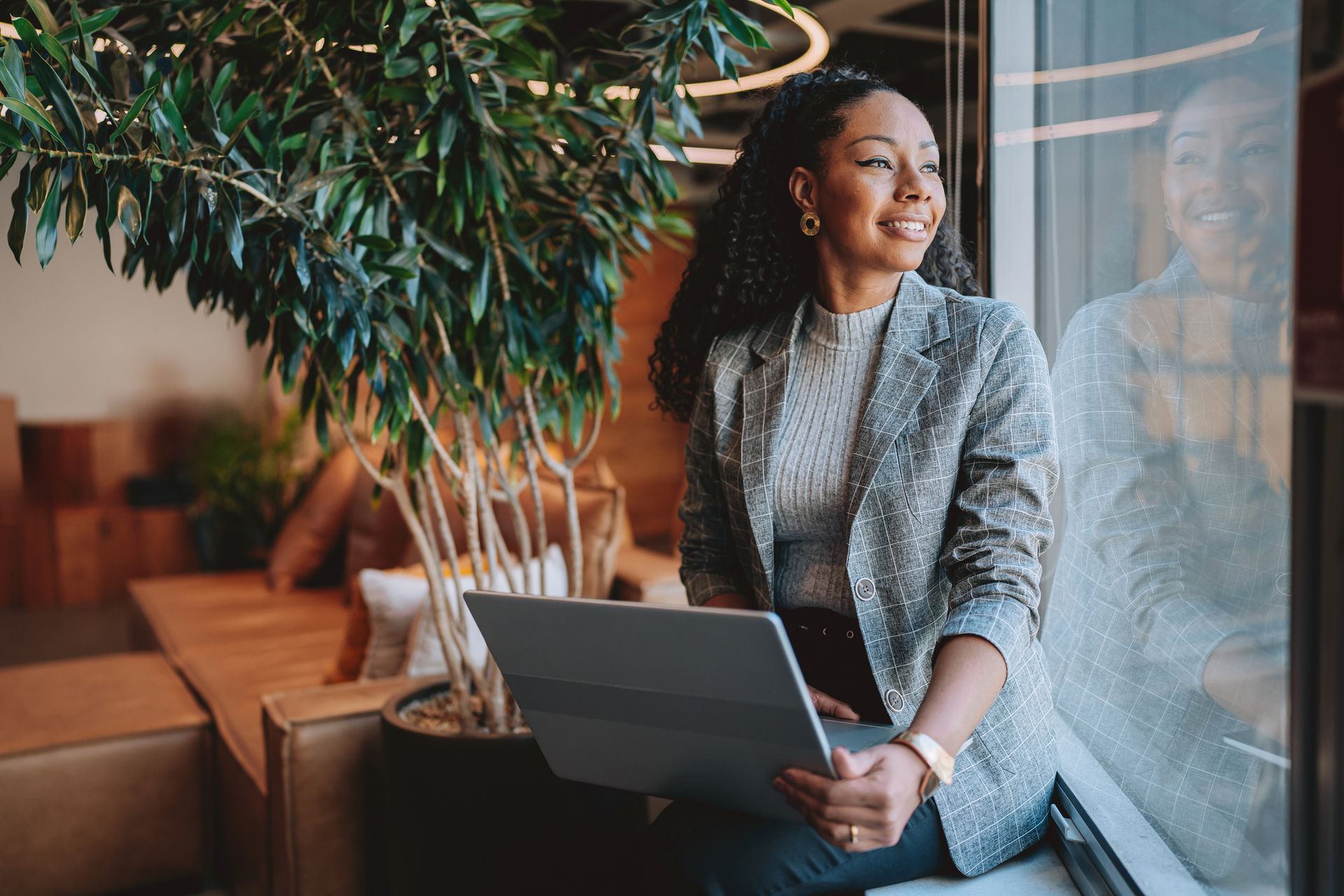 Woman in a blazer smiles, looking out a window while holding a laptop; next to a plant in an office.