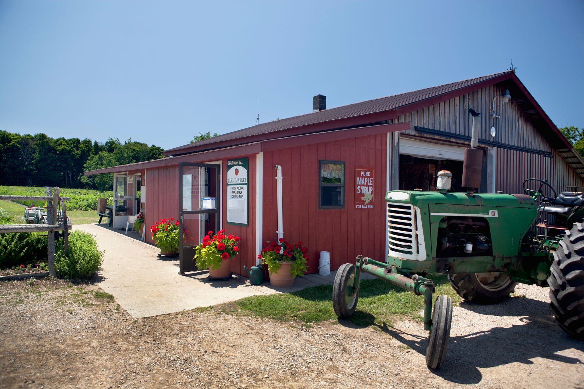 Red farm stand with potted flowers, concrete path, and green tractor parked outside on a sunny day.