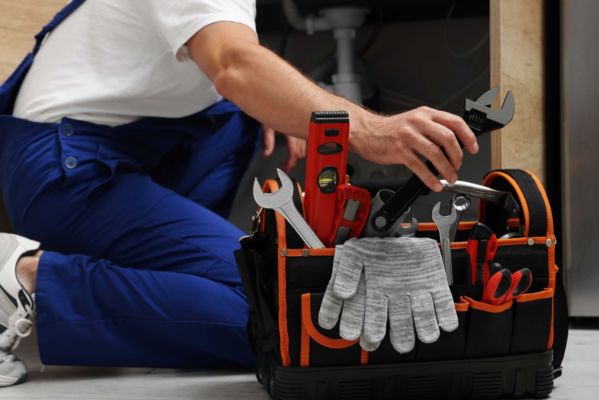 A plumber is kneeling down next to a tool bag filled with tools.