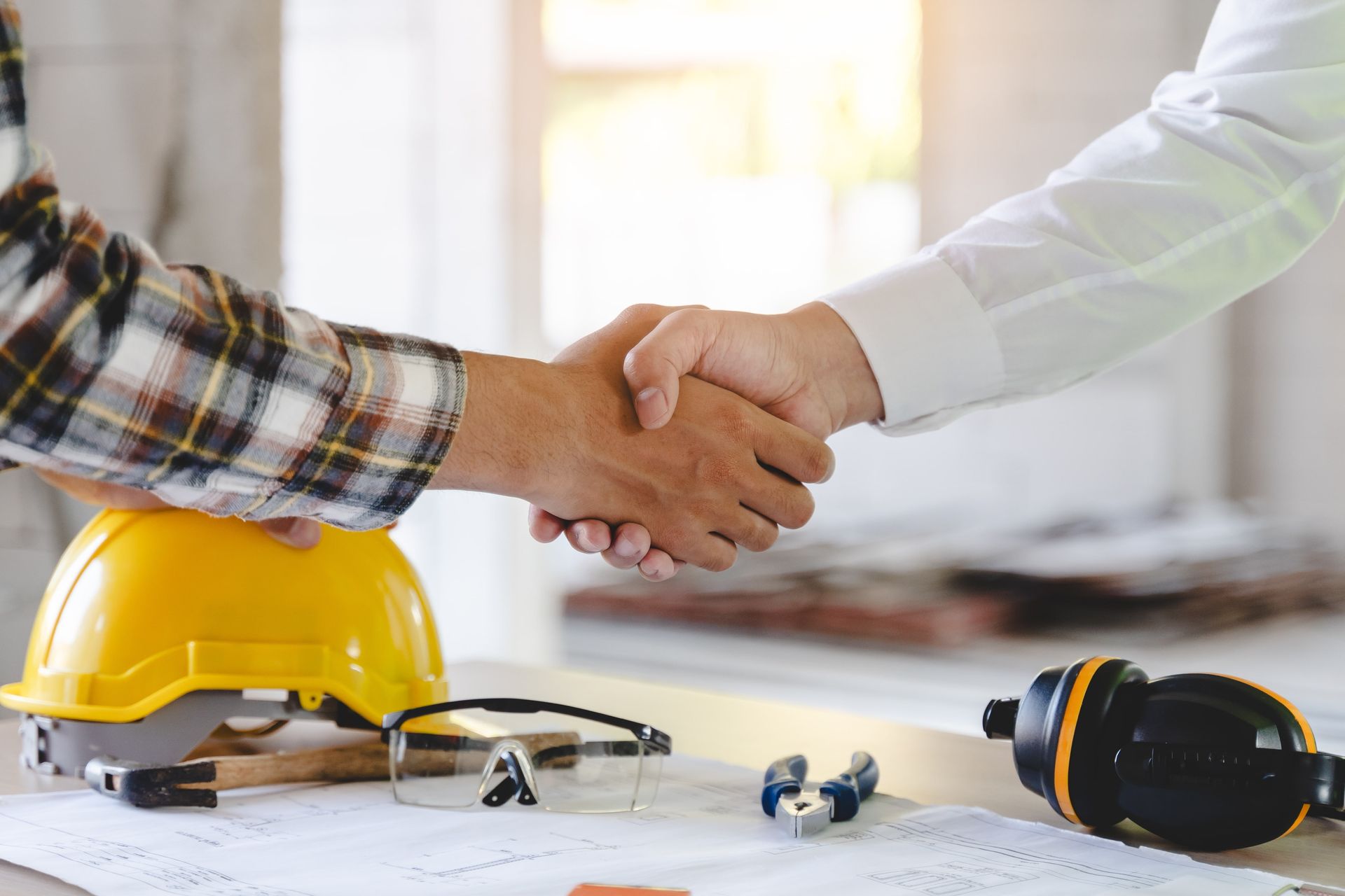 Two men are shaking hands over a table with a hard hat on it.