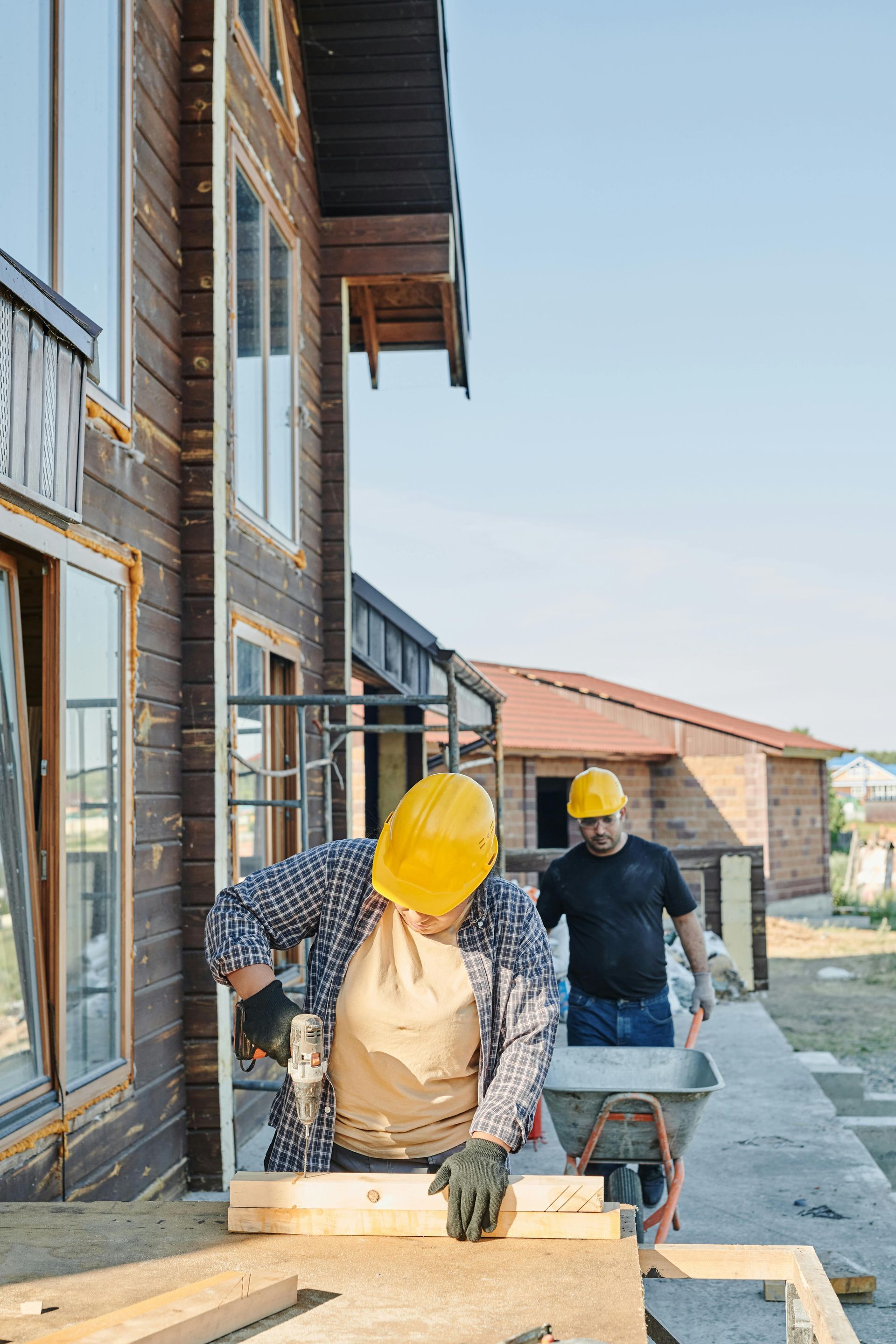 Two construction workers in yellow helmets working on a wood structure outside a brown building.