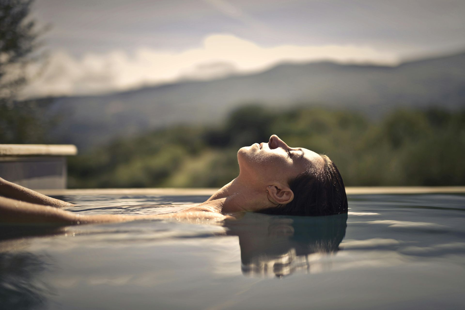 Person relaxing in a pool, head tilted back, mountain view in background.