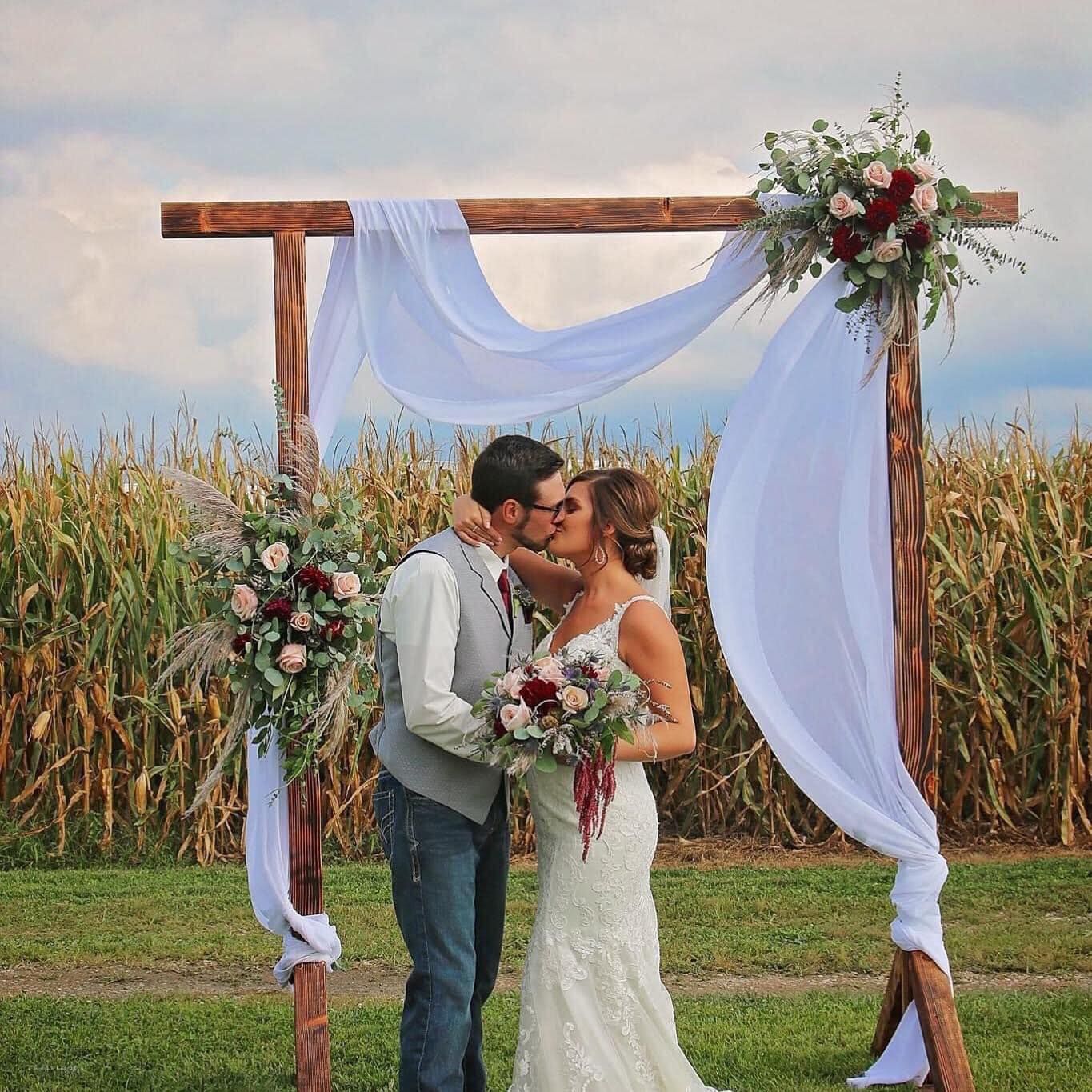 A bride and groom are kissing under a wooden arch in front of a corn field.