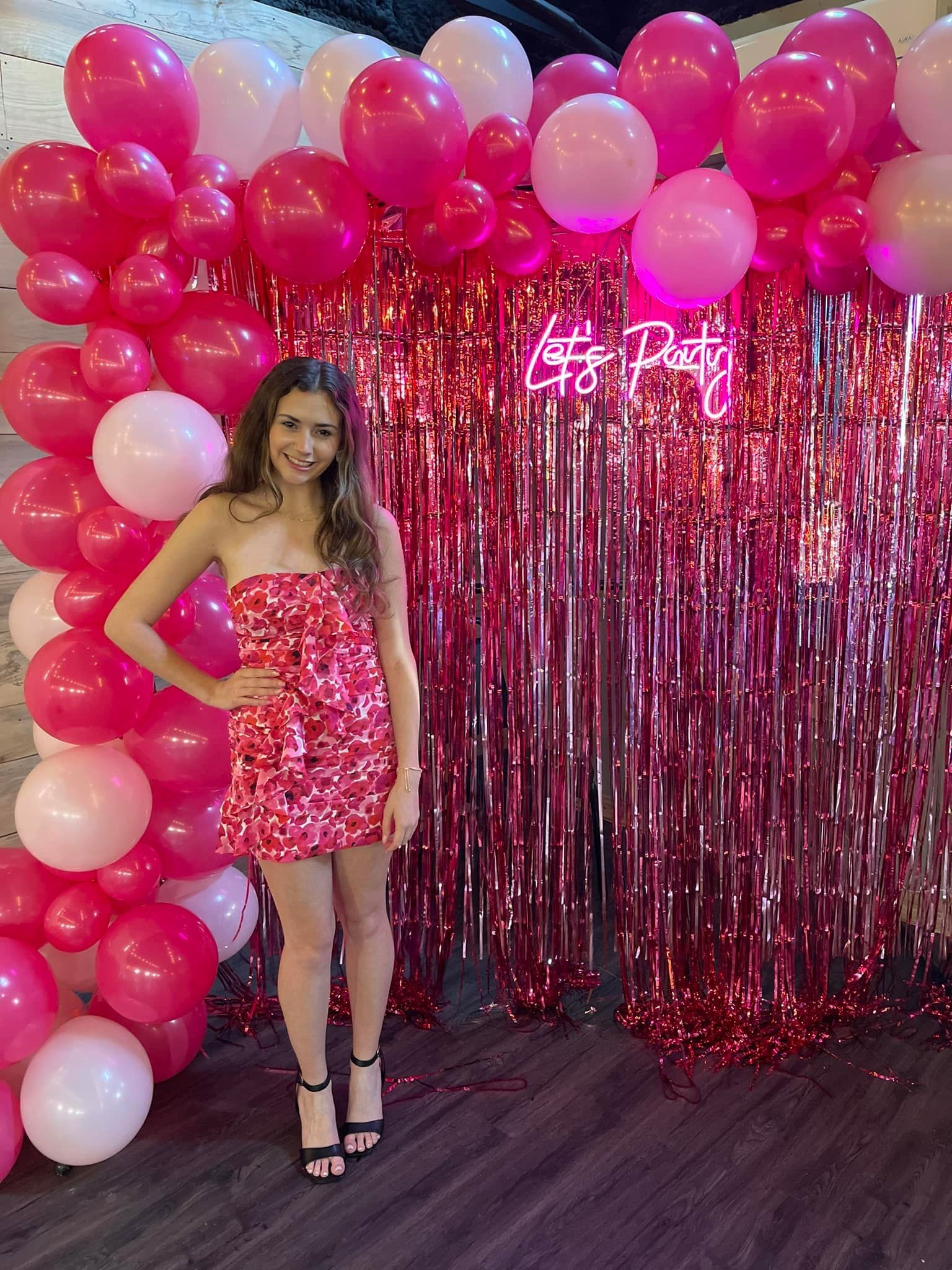 A woman in a pink dress is standing in front of a wall of pink and white balloons.