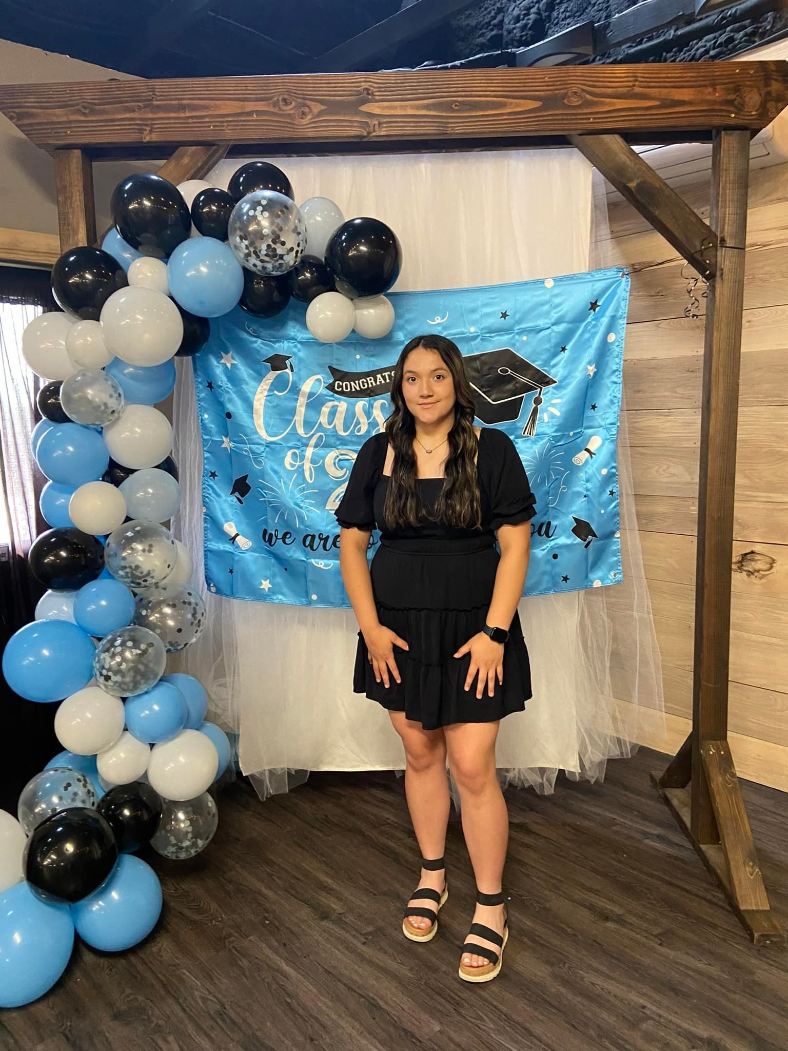 A woman in a black dress is standing in front of a graduation banner and balloons.