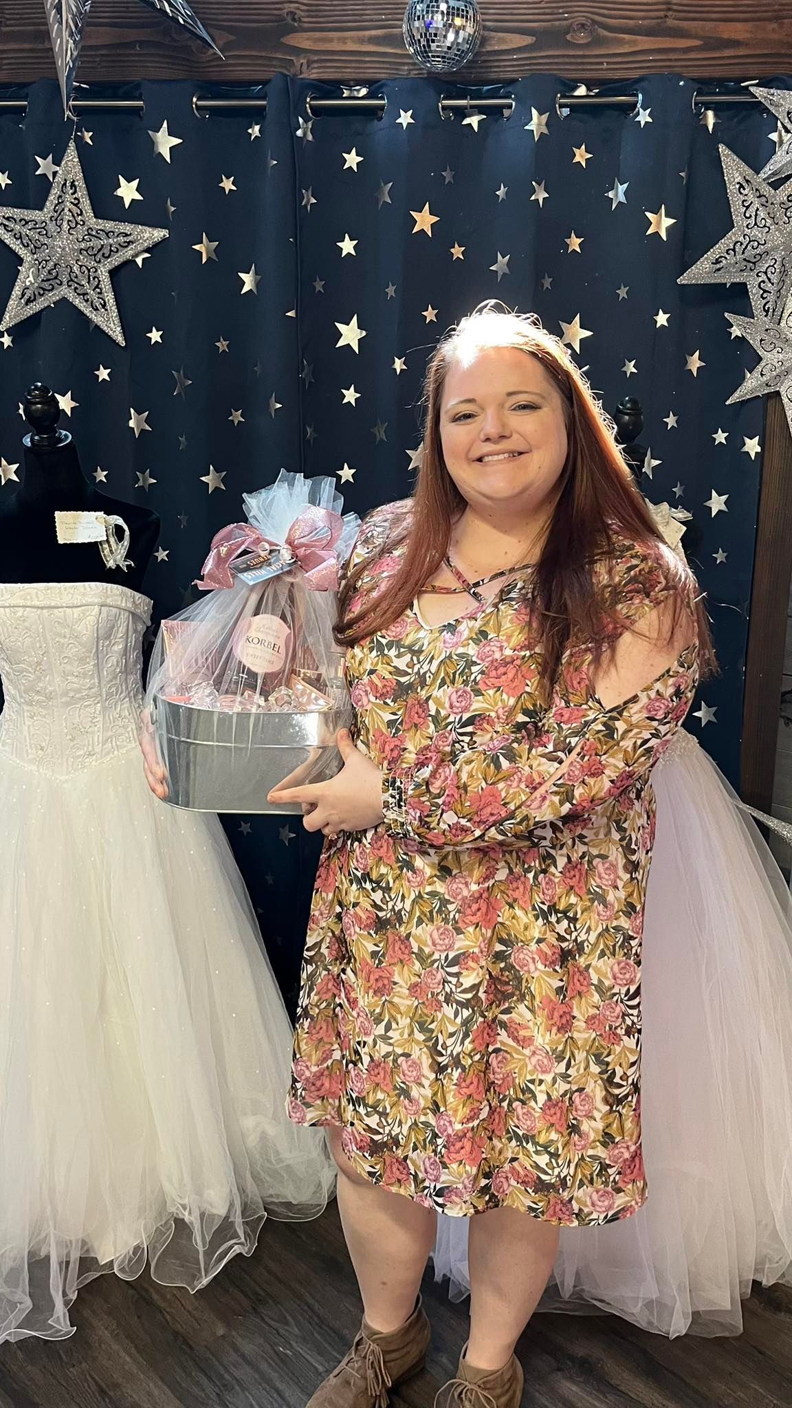 A woman in a dress is holding a gift basket in front of a wedding dress.