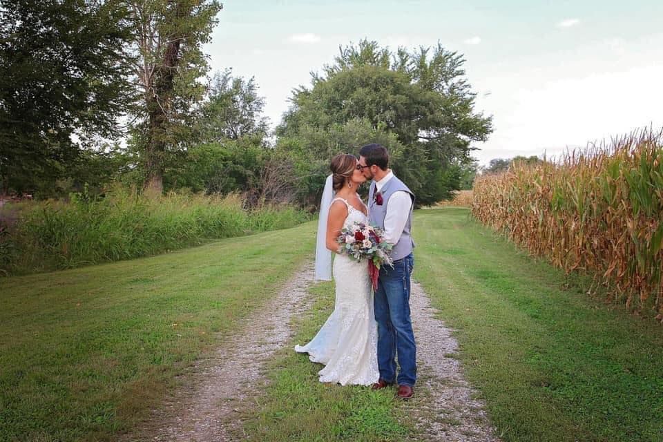 A bride and groom are kissing on a dirt road in front of a corn field.