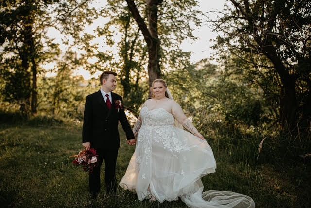 A bride and groom are holding hands in a field . the bride is wearing a plus size wedding dress.