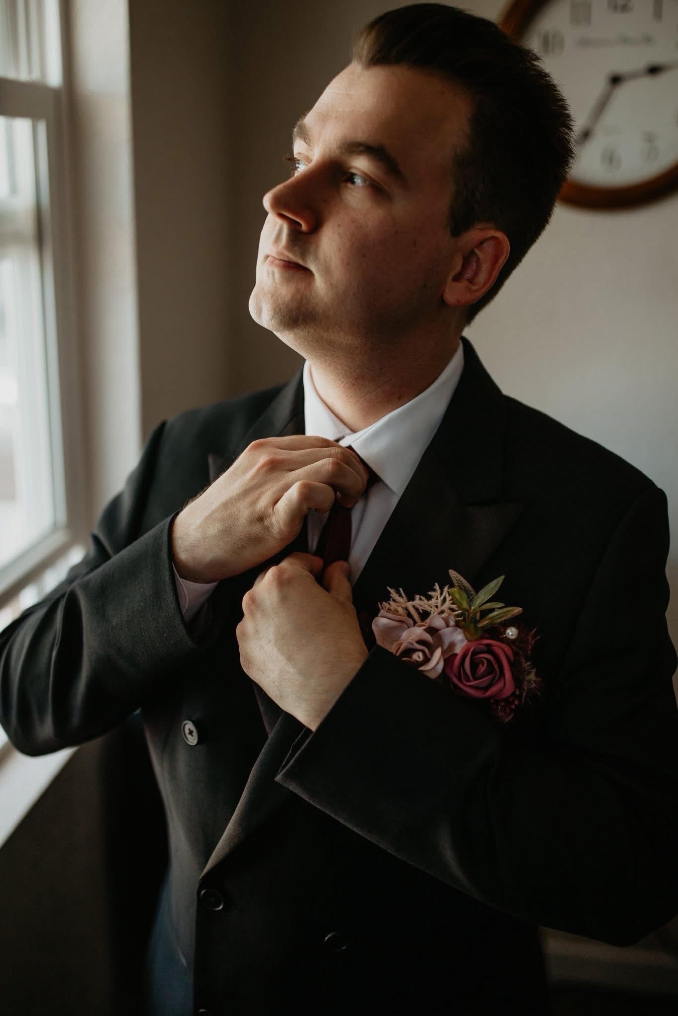 A man in a suit is adjusting his tie while looking out a window.