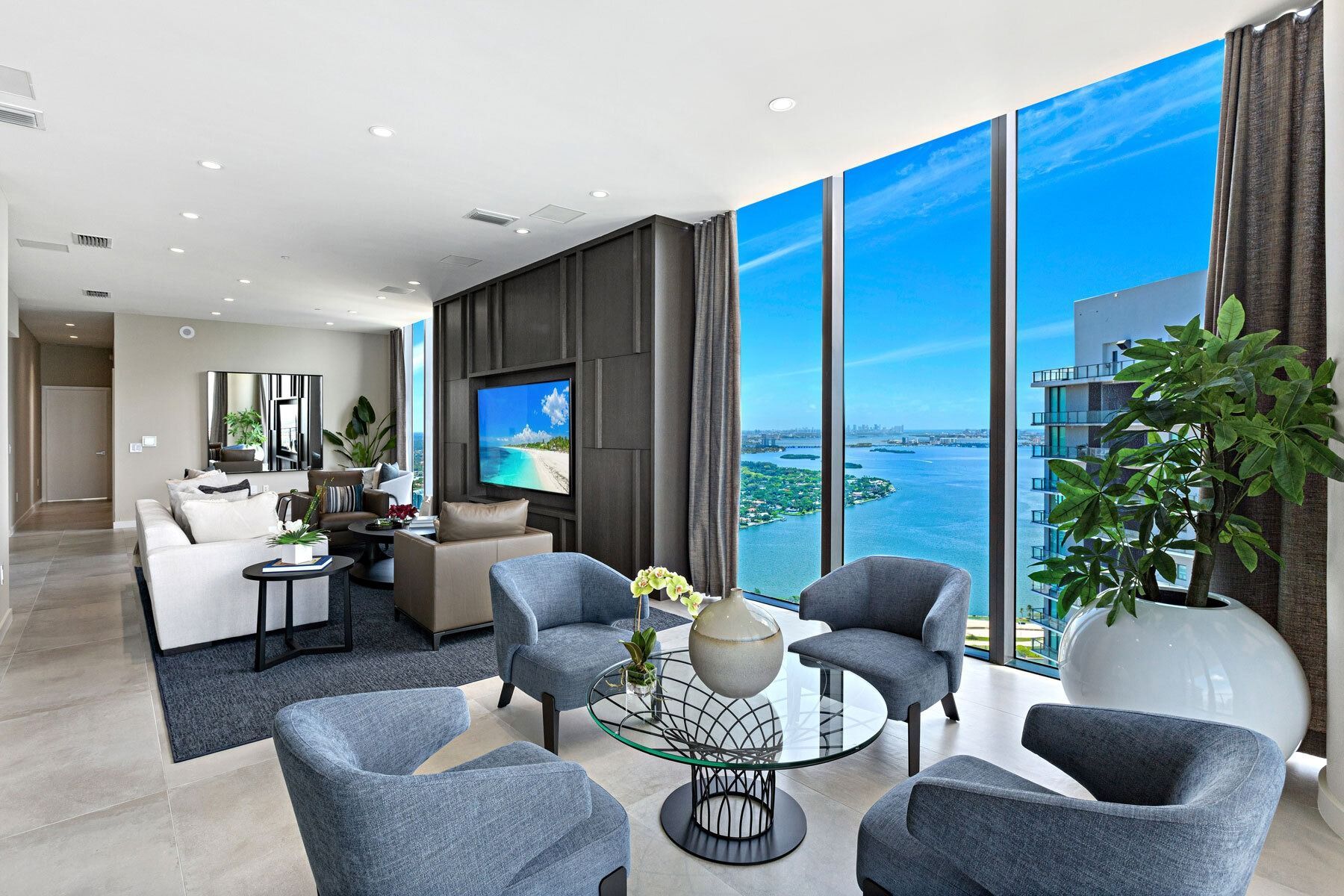 Living room with large windows overlooking a blue ocean.  Gray chairs and sofa, TV, and indoor plant.