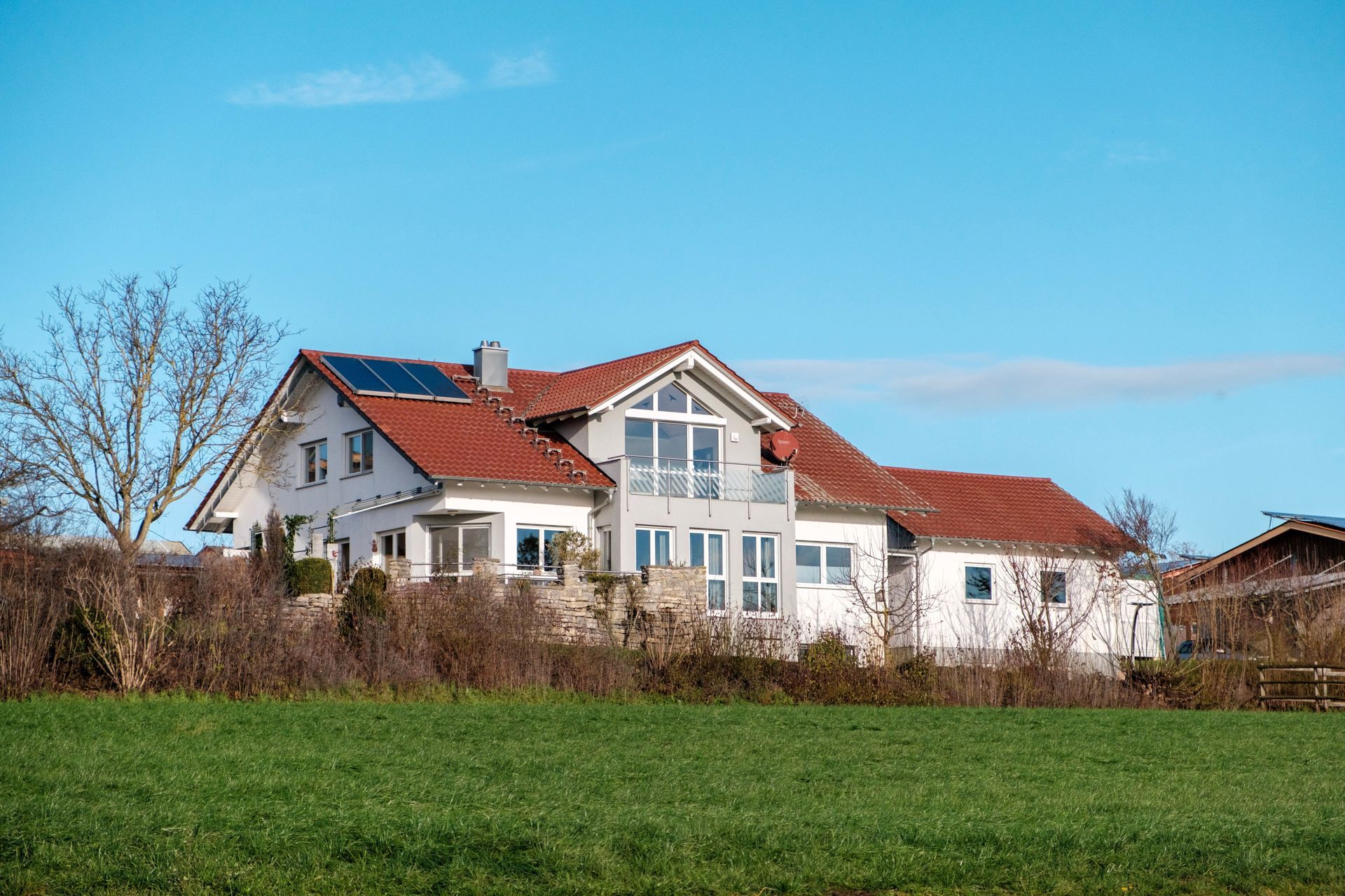 House with white walls, red roof, and solar panels on a grassy hill under a blue sky.
