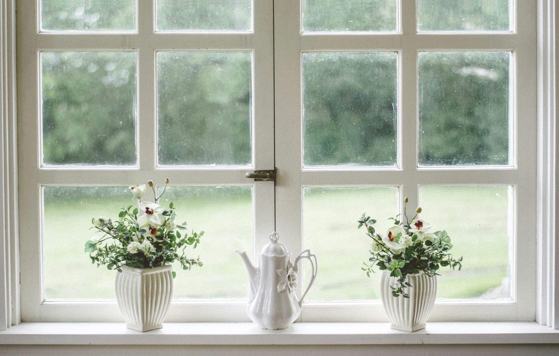 White framed window with a teapot and potted flowers. Green foliage can be seen outside the glass.