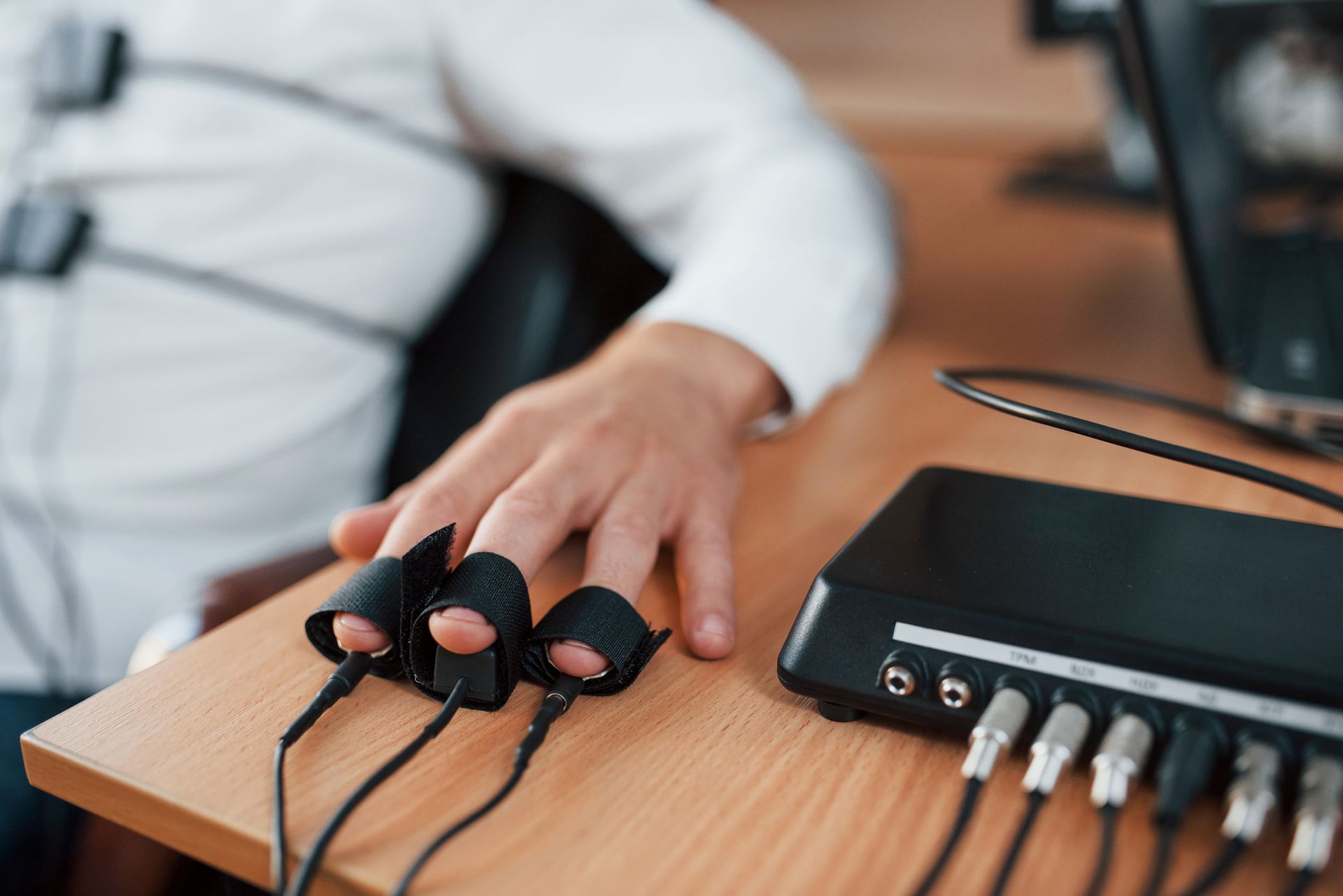 A person's hand with sensors attached to their fingers, connected to a black device on a wooden table. The setting appears to be an office.