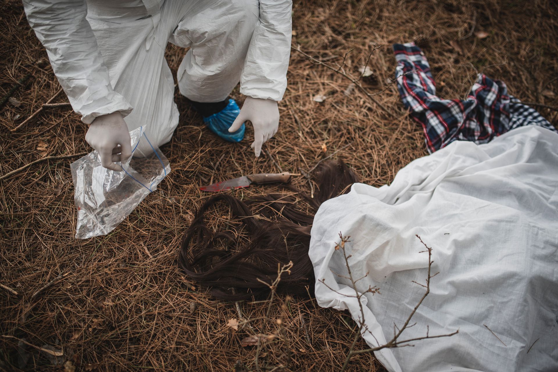 A crime scene with a body covered by a white sheet. A person in a white suit examines evidence on the ground.