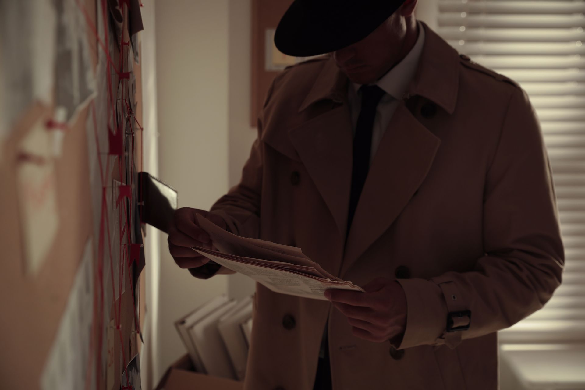 A detective in a trench coat and fedora examines a bloody crime scene wall, holding papers.