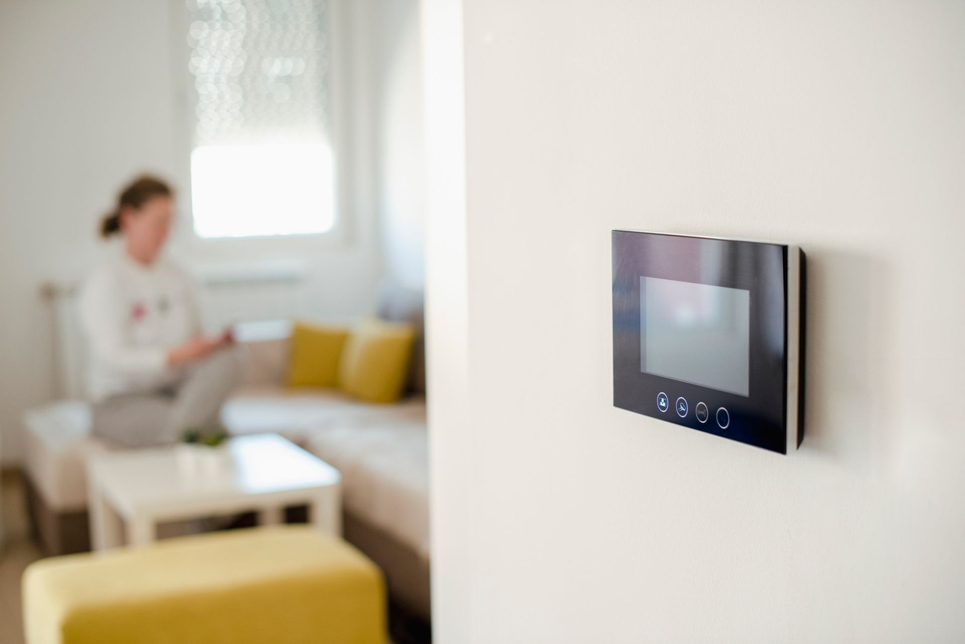 Black intercom device mounted on a white wall; a person sits on a couch in the blurred background.