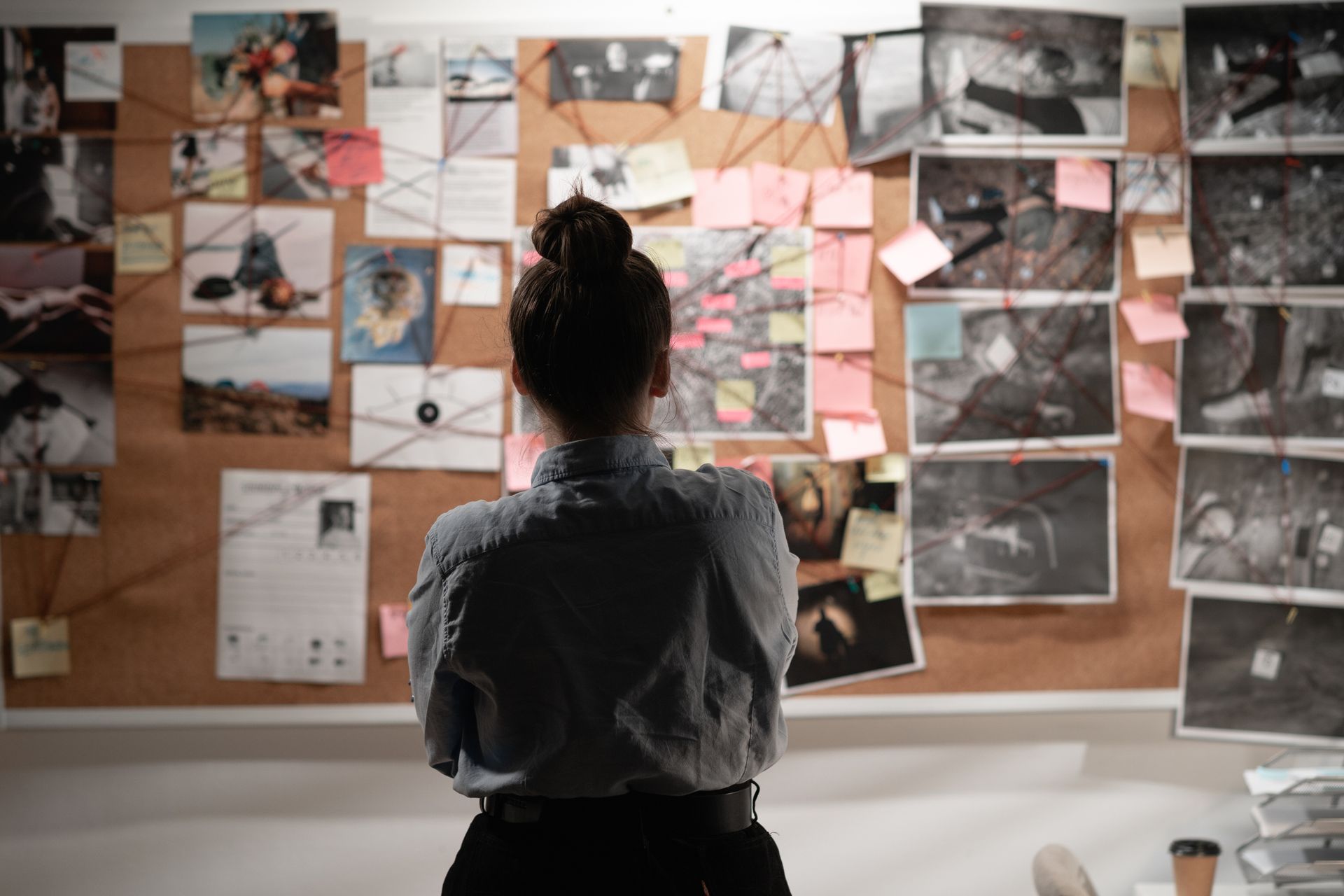 A woman stands before a corkboard covered in photos, notes, and string, seemingly analyzing the information; an investigator at work.