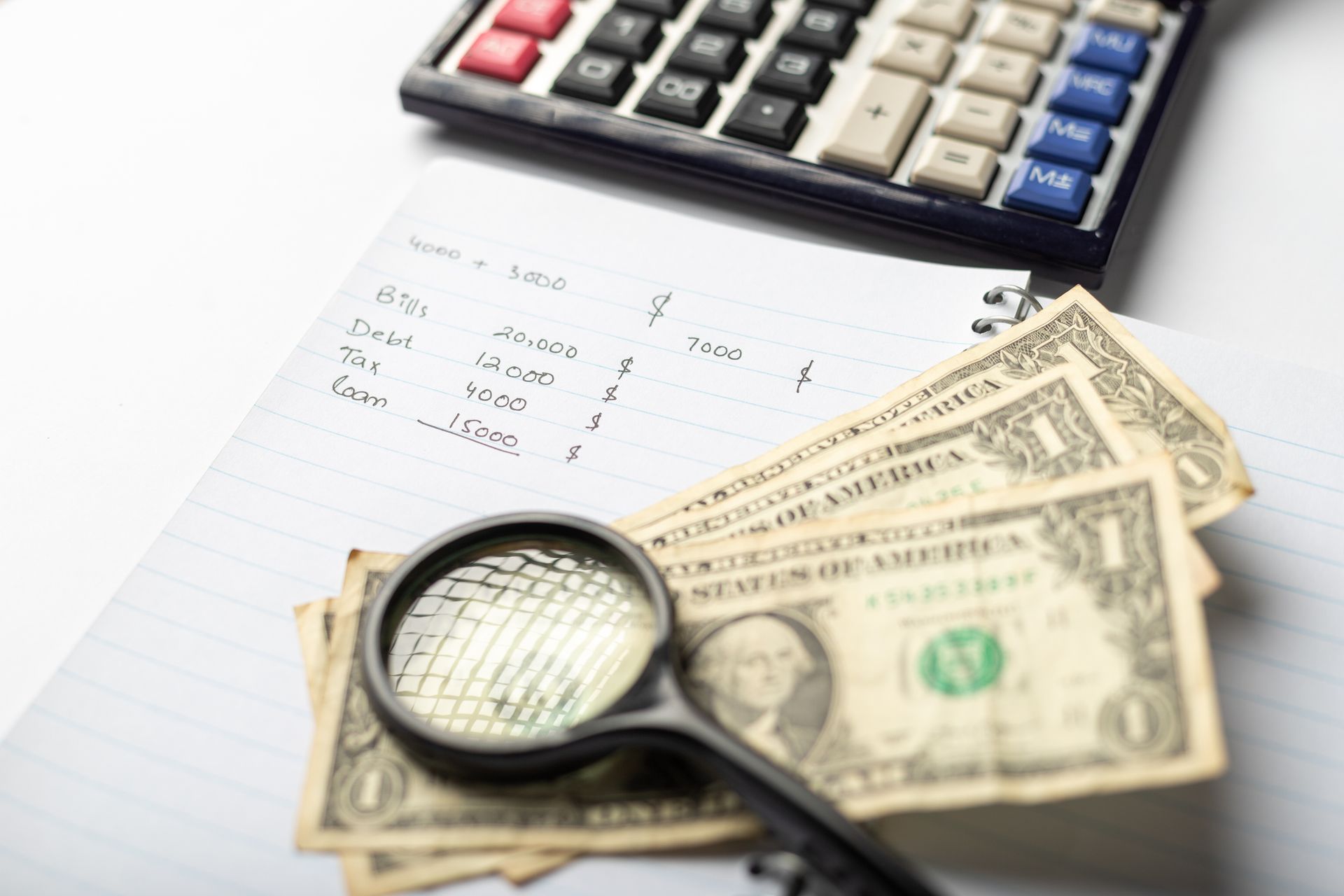 Calculator, handwritten list, magnifying glass, and dollar bills on white paper, likely for budgeting or financial review.