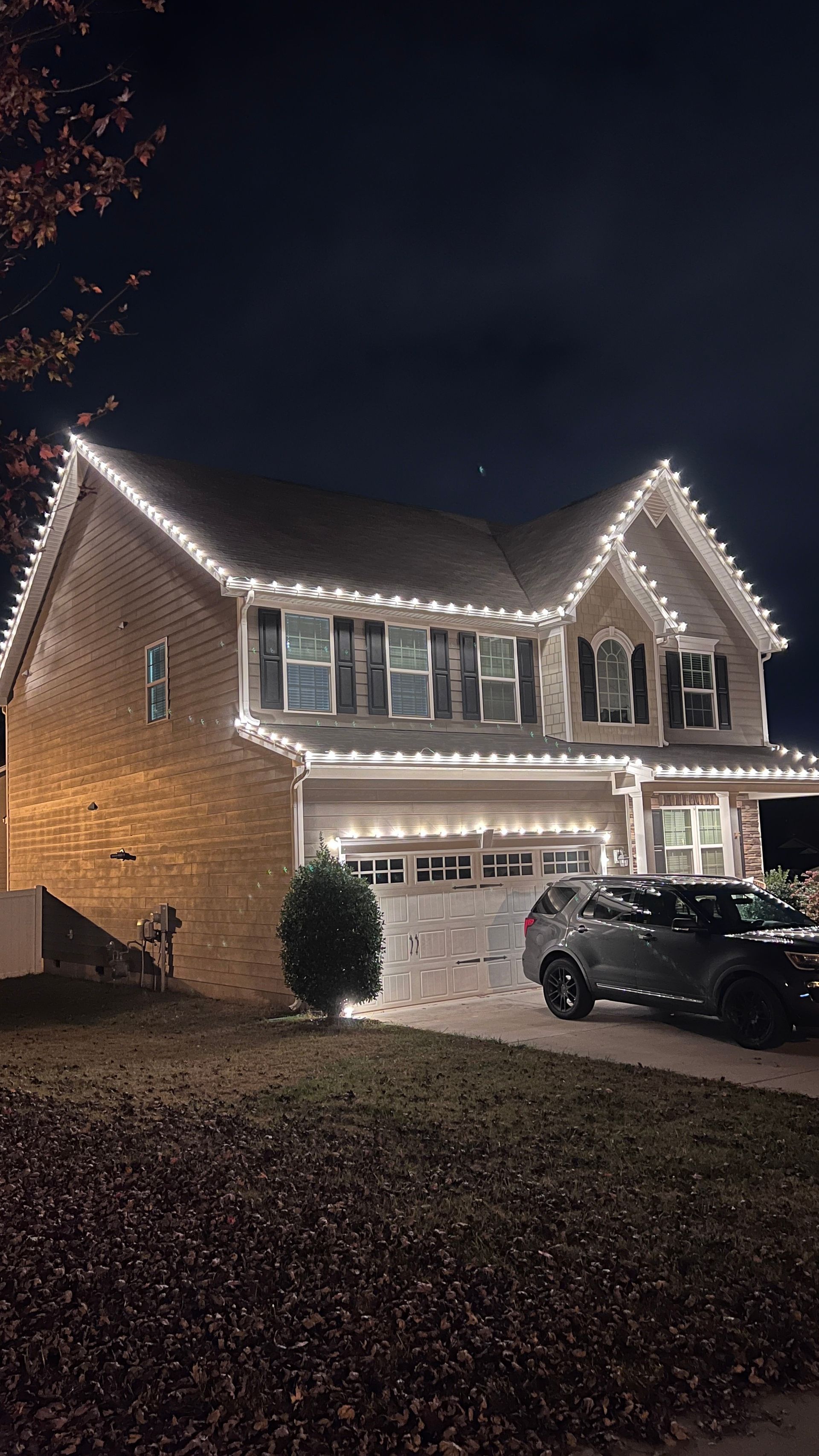 House at night with white Christmas lights outlining the roof and windows. A car is parked in the driveway.