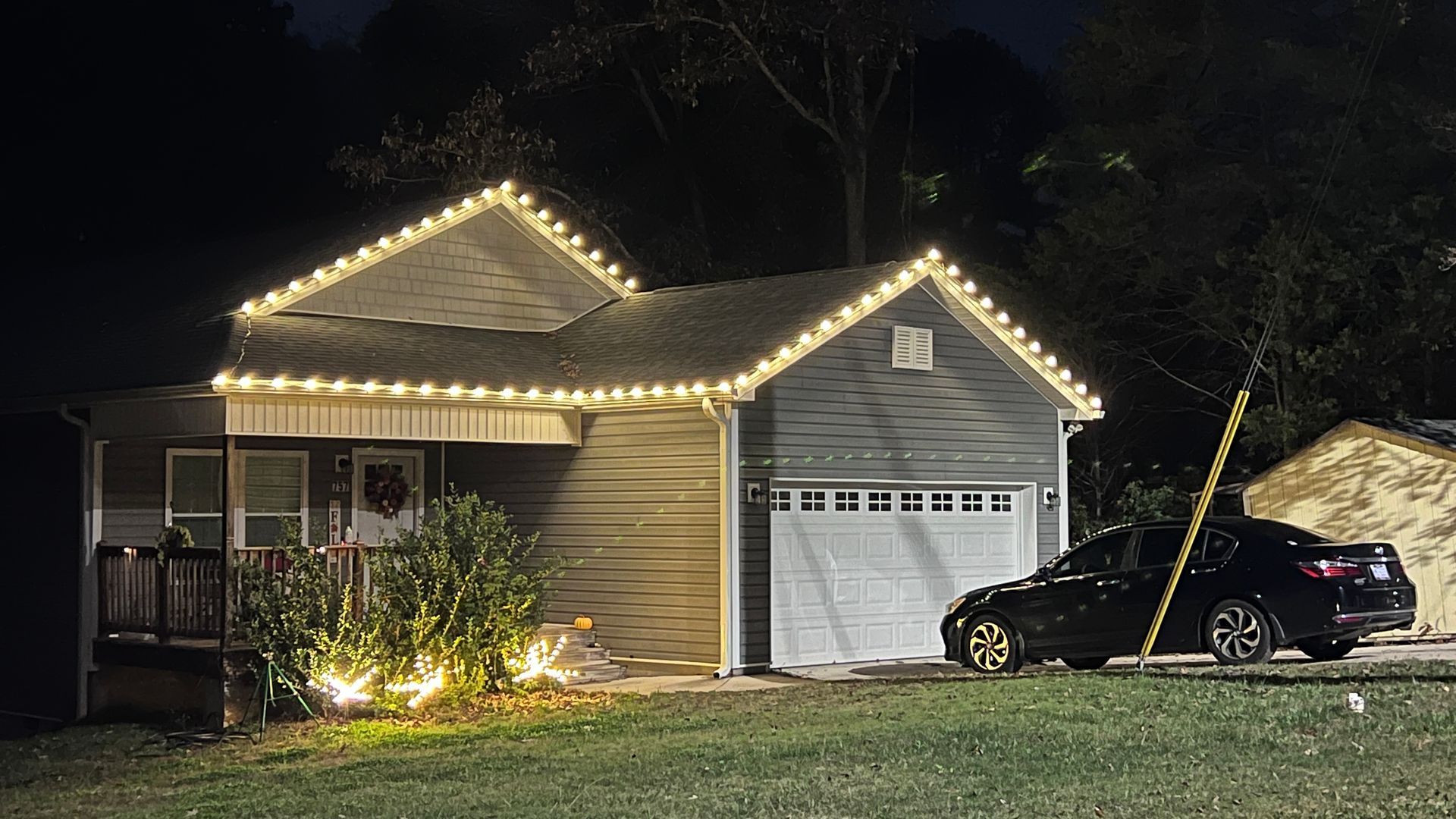 House at night with roofline Christmas lights, lit bushes, and car parked in front of garage.