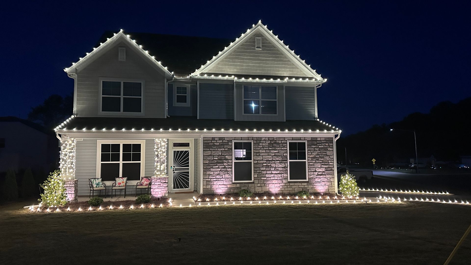 Two-story house illuminated with white Christmas lights along the roofline, windows, and lawn. Pink spotlights at the base.