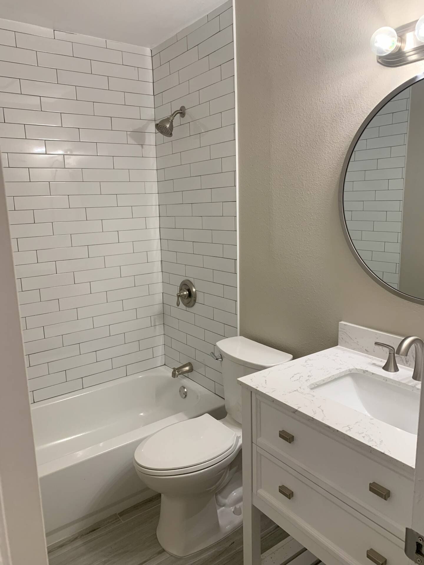 White bathroom with tub and shower, white tile, white vanity, round mirror.