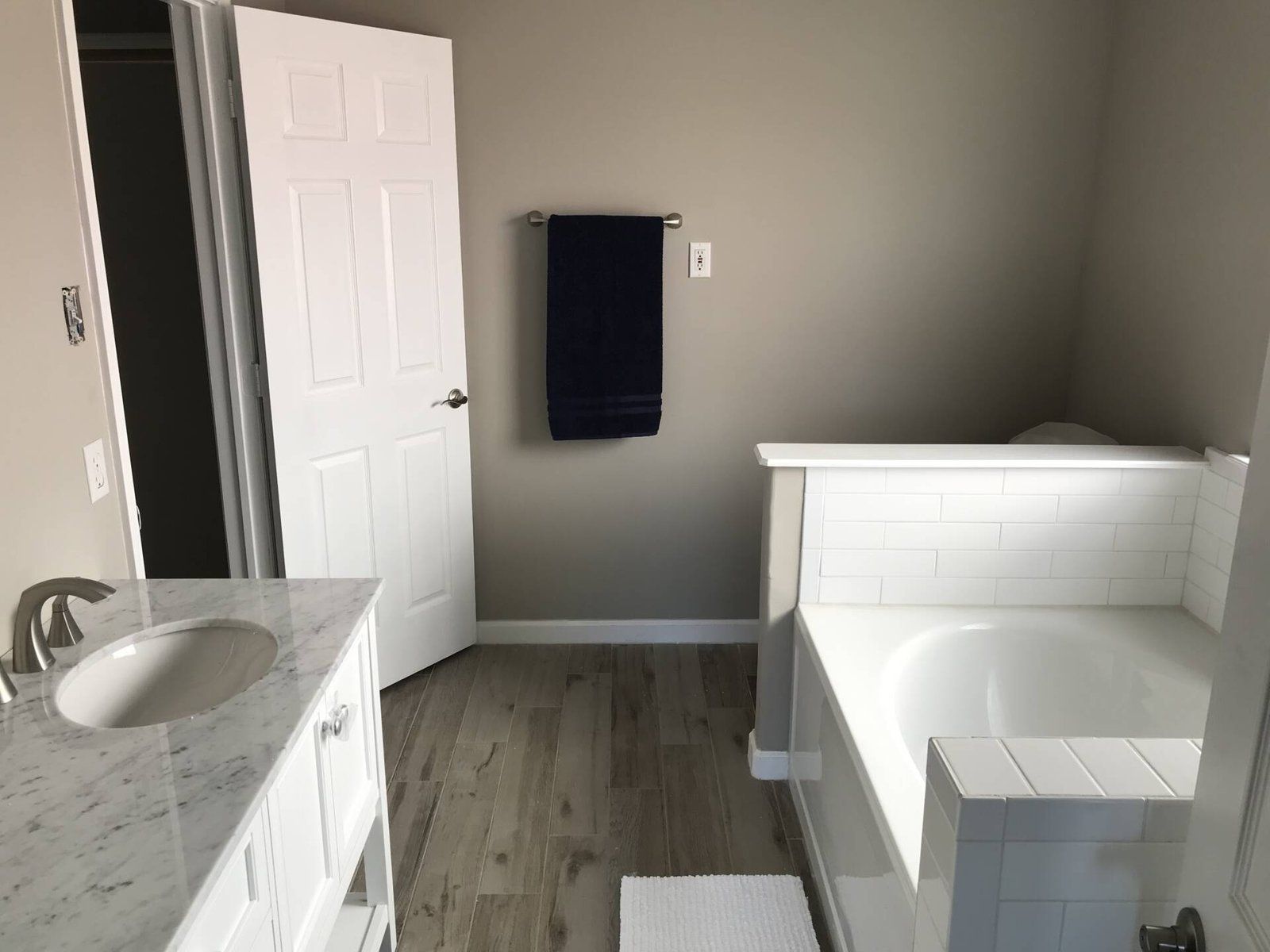 Bathroom with a white bathtub, vanity, and door; gray walls; wood-look floor; black towel.