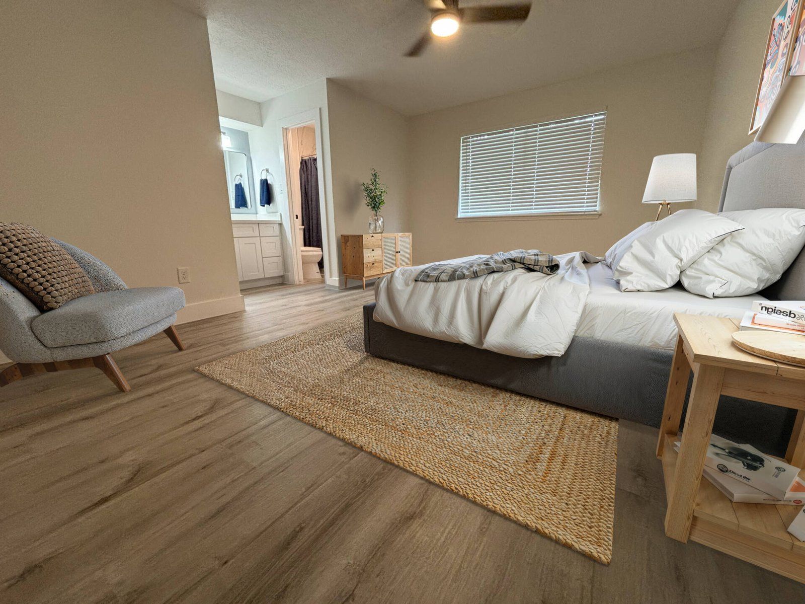 Bedroom with bed, armchair, rug, and open doorway to bathroom. Beige walls and light wood flooring.