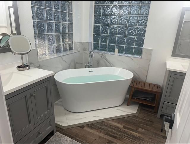 Modern bathroom with a white tub, gray vanities, glass block window, and a wooden bench.