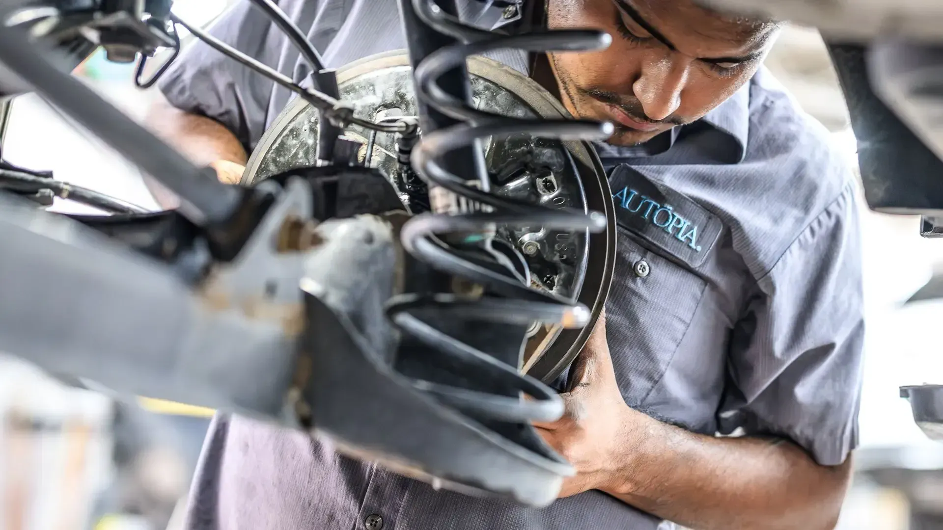 Mechanic working on car's undercarriage; close up. He wears a grey shirt and intently examines brakes | Autopia Auto Repair - Bloomington