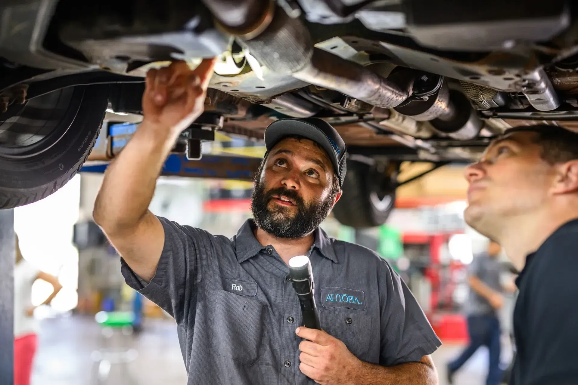 Mechanic pointing under car, explaining to another person in a shop | Autopia Auto Repair - Bloomington