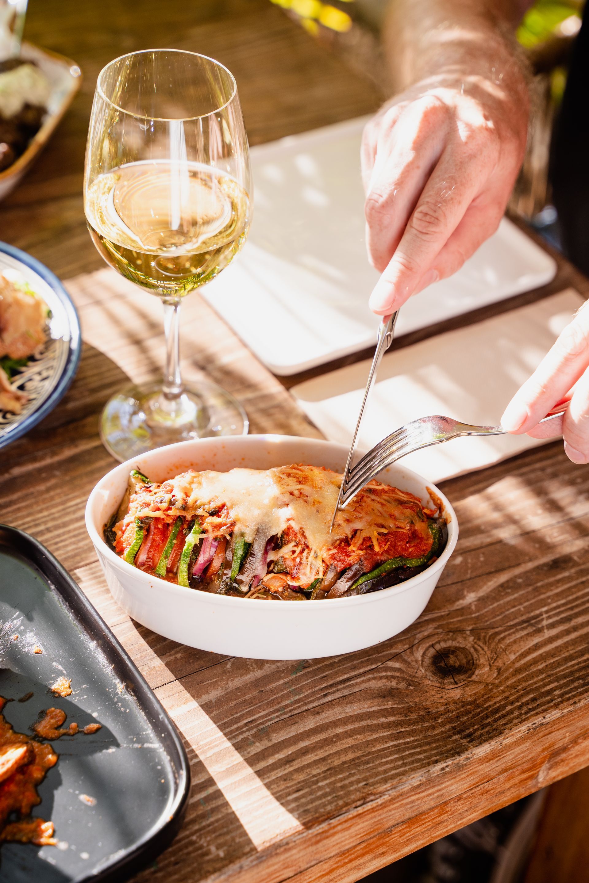 A person is cutting a piece of food with a fork on a table with a glass of wine.