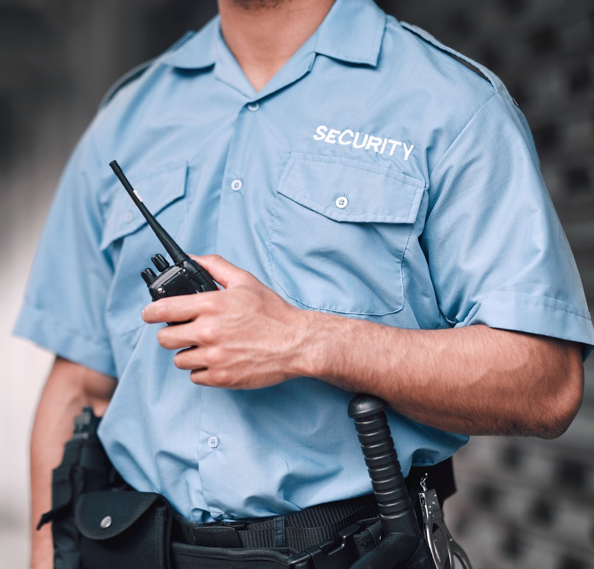 Guardia de seguridad con uniforme azul, con radio en la mano. De pie, con una porra atada al cinturón.