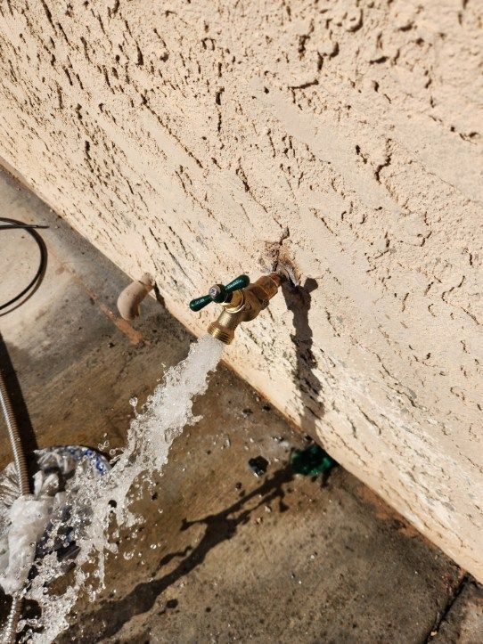 Water gushing from an outdoor faucet attached to a stucco wall, creating a stream on the concrete.