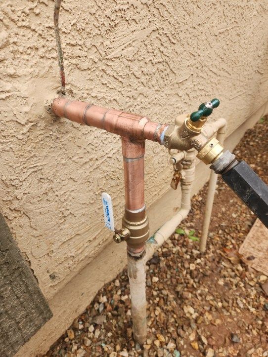 Copper and brass outdoor water pipes and valves against a stucco wall, with a connected hose.