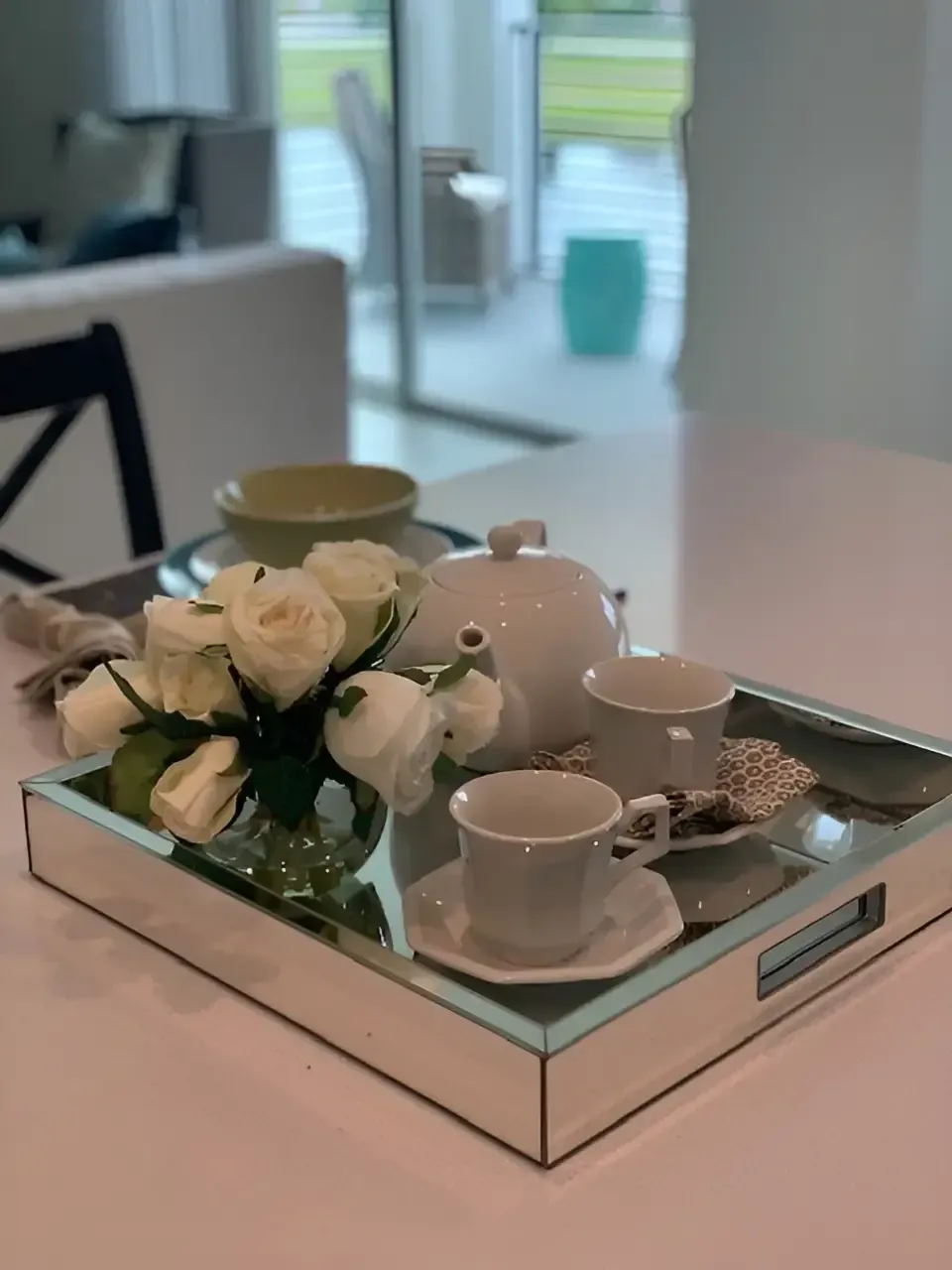 A white tea set and a small vase of white roses on a mirrored tray atop a table in a bright, modern room.