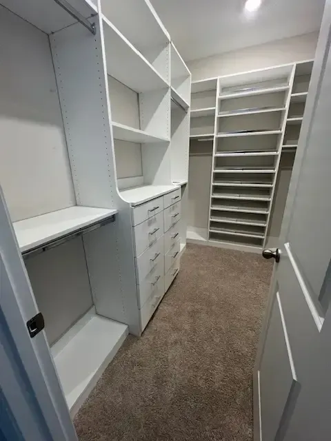 A walk-in closet featuring white built-in shelving, a dresser unit with drawers, and carpeted flooring.