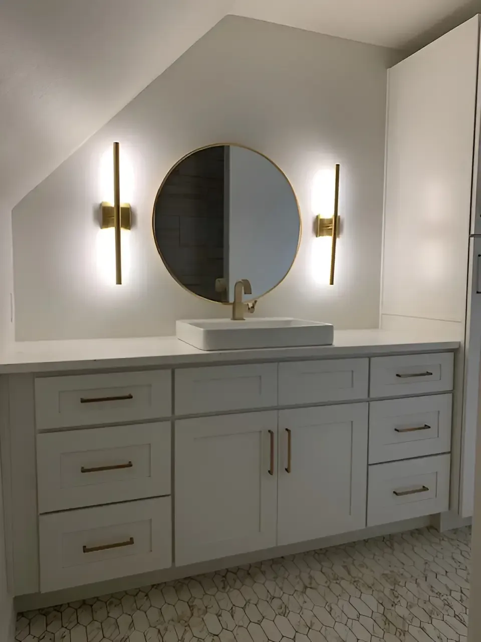 A bathroom vanity with white cabinets, gold hardware, a round mirror, and two wall-mounted light fixtures.