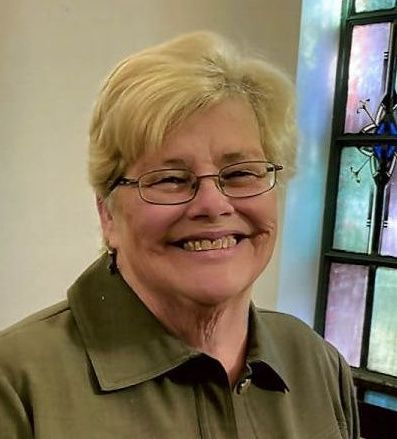 A woman wearing glasses is smiling in front of a stained glass window.