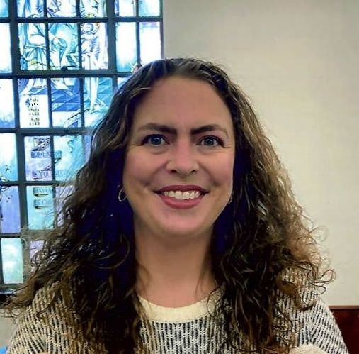 A woman with long curly hair is smiling in front of a stained glass window.
