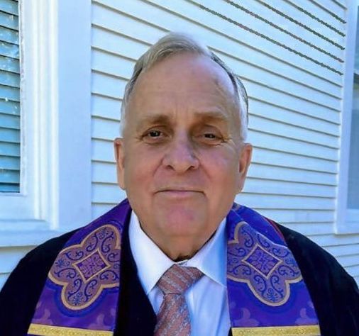 A man wearing a purple stole and tie stands in front of a white house