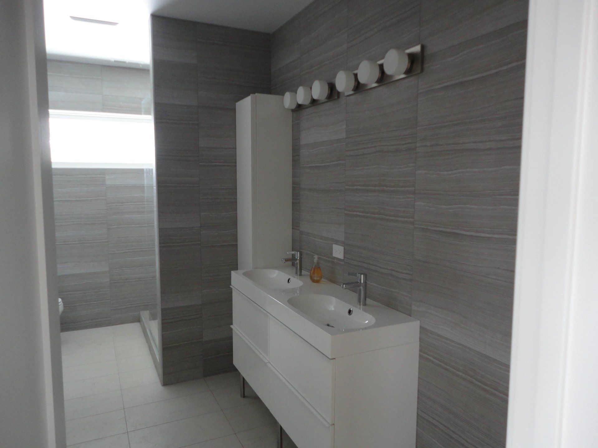 Modern gray-tiled bathroom with a white double vanity, cabinet, and light fixture. Shower visible in the background.