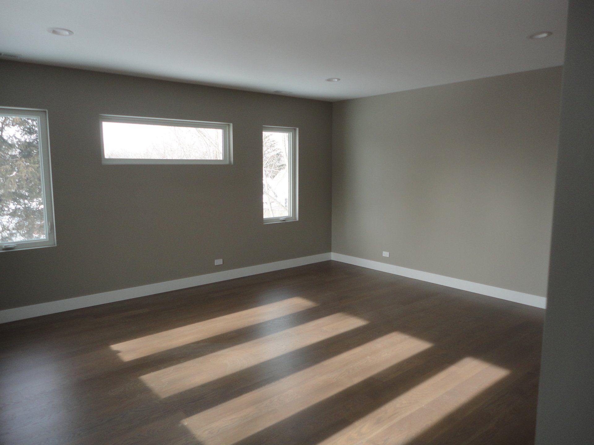Empty room with brown hardwood floor, neutral walls, and sunlight streaming in windows.