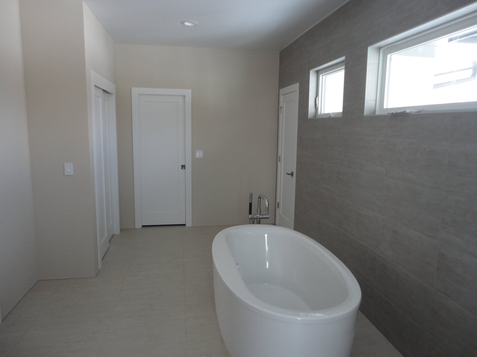 Modern bathroom with a freestanding tub, gray accent wall, two windows, and white doors.