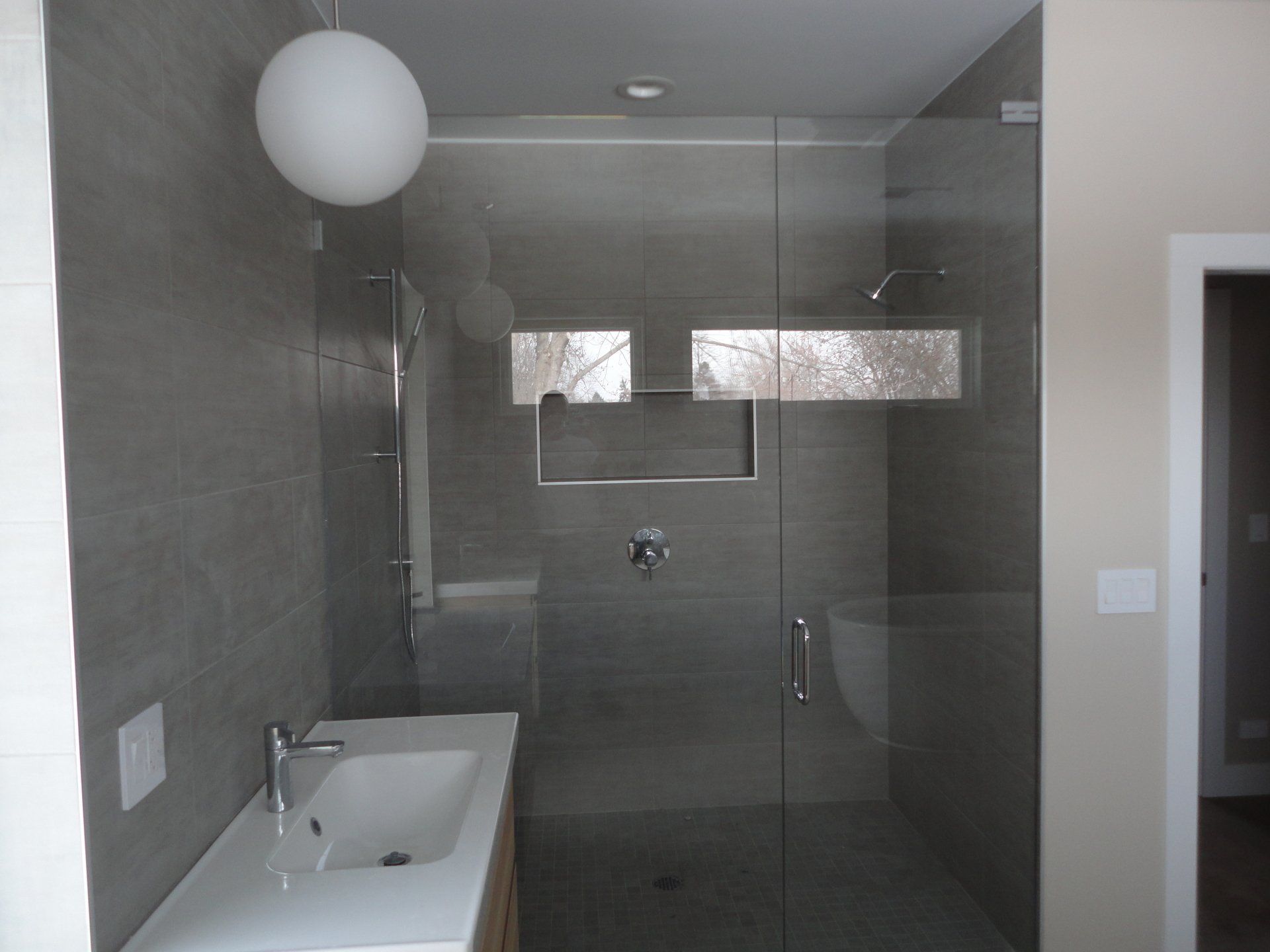 Modern bathroom with a glass shower, gray tile, white sink, and a hanging globe light.